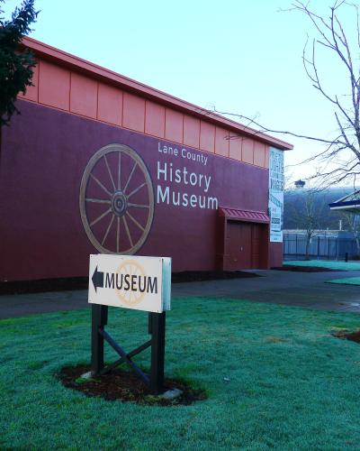 The image shows the exterior of a building with a large, painted wooden wagon wheel on its reddish-brown wall, a white sign in the foreground reading "MUSEUM" with an arrow, and a blue trash can on the grass.