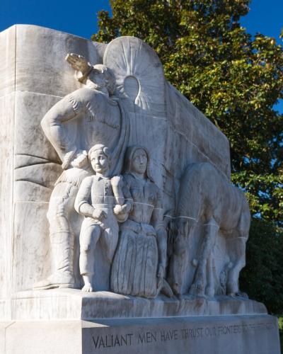 A close-up view of a large white marble bas-relief sculpture depicting pioneers and a covered wagon, with the inscription "VALIANT MEN HAVE THRUST OUR FRONTIERS TO THE SETTING SUN" on the base, situated next to a marble building.