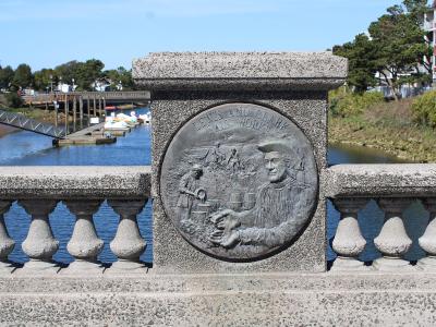 A close-up of a bronze commemorative plaque mounted on a concrete bridge railing, depicting figures and the text "LEWIS AND CLARK SALT WORKS" with the date "1806", overlooking a river with paddle boats and a dock.
