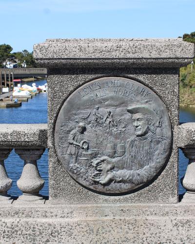A close-up of a bronze commemorative plaque mounted on a concrete bridge railing, depicting figures and the text "LEWIS AND CLARK SALT WORKS" with the date "1806", overlooking a river with paddle boats and a dock.