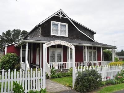 The image shows a single-story, dark red wooden cottage with white trim, a white picket fence, and a sign above the front porch entrance that reads "BUTTERFIELD COTTAGE".