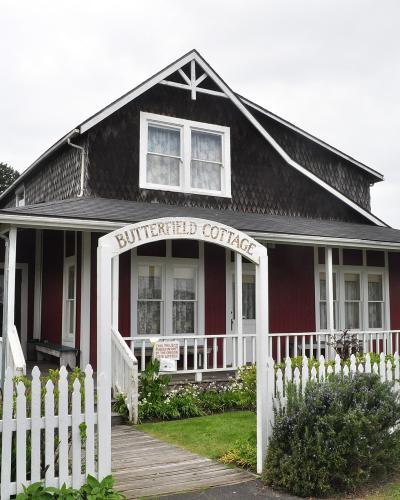 The image shows a single-story, dark red wooden cottage with white trim, a white picket fence, and a sign above the front porch entrance that reads "BUTTERFIELD COTTAGE".