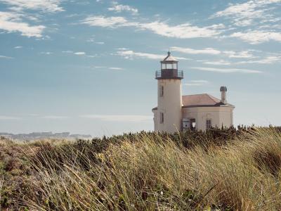 A white lighthouse with an attached keeper's house stands on a grassy dune overlooking the ocean under a partly cloudy sky.