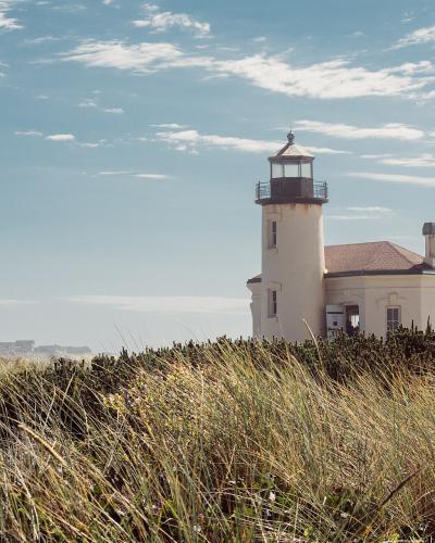 A white lighthouse with an attached keeper's house stands on a grassy dune overlooking the ocean under a partly cloudy sky.