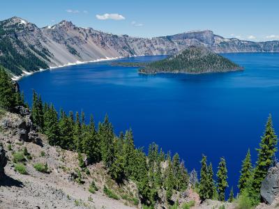 A landscape photograph showing a deep blue lake surrounded by steep, rocky volcanic cliffs, with a prominent, cone-shaped, tree-covered island in the center of the lake.