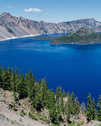 A landscape photograph showing a deep blue lake surrounded by steep, rocky volcanic cliffs, with a prominent, cone-shaped, tree-covered island in the center of the lake.