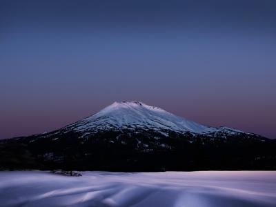A snow-covered stratovolcano at twilight, with a foreground of snow and a few conifer trees on the left.