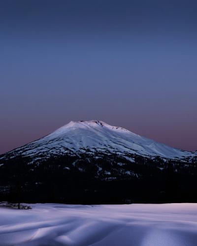 A snow-covered stratovolcano at twilight, with a foreground of snow and a few conifer trees on the left.