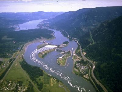 An aerial photograph of a large hydroelectric dam complex situated on a wide river within a steep, forested gorge, featuring a prominent navigation lock structure and spillway.