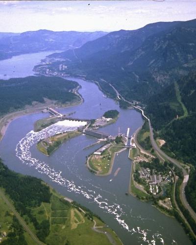 An aerial photograph of a large hydroelectric dam complex situated on a wide river within a steep, forested gorge, featuring a prominent navigation lock structure and spillway.