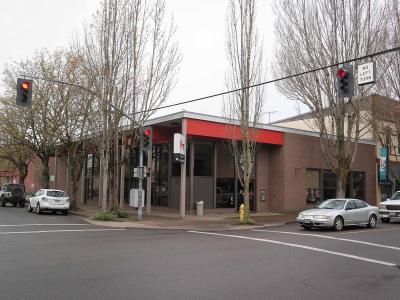 A street-level view of a single-story brick commercial building with a red fascia and large glass windows, situated at an intersection with traffic lights and bare trees.