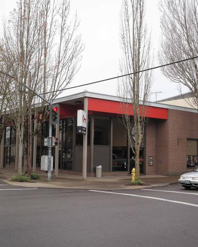 A street-level view of a single-story brick commercial building with a red fascia and large glass windows, situated at an intersection with traffic lights and bare trees.