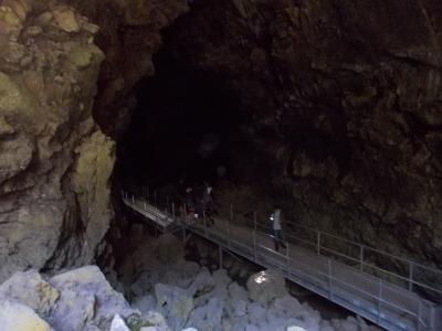 A view inside a dark, natural lava tube cave featuring a metal walkway with railings, where several people are walking; the rock walls are rough and illuminated by artificial light.