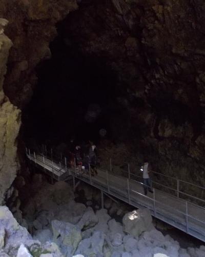 A view inside a dark, natural lava tube cave featuring a metal walkway with railings, where several people are walking; the rock walls are rough and illuminated by artificial light.