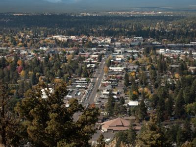 A high-angle panoramic view of a city (Bend, Oregon) surrounded by forests and distant mountains, taken from an elevated vantage point with sagebrush and trees in the foreground.