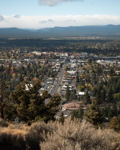 A high-angle panoramic view of a city (Bend, Oregon) surrounded by forests and distant mountains, taken from an elevated vantage point with sagebrush and trees in the foreground.