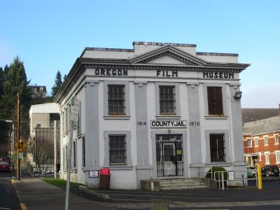 A white, two-story building with barred windows and signage reading "OREGON FILM MUSEUM" across the top and "COUNTY JAIL" with dates "1914" and "1976" above the entrance.