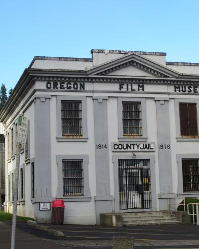 A white, two-story building with barred windows and signage reading "OREGON FILM MUSEUM" across the top and "COUNTY JAIL" with dates "1914" and "1976" above the entrance.
