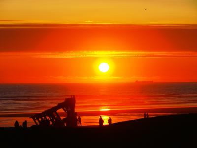 A silhouette of a large, rusted shipwreck on a sandy beach at sunset, with several people standing near the wreck and a cargo ship visible on the horizon.