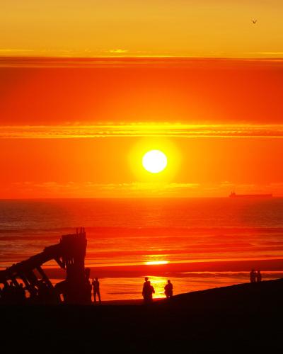 A silhouette of a large, rusted shipwreck on a sandy beach at sunset, with several people standing near the wreck and a cargo ship visible on the horizon.