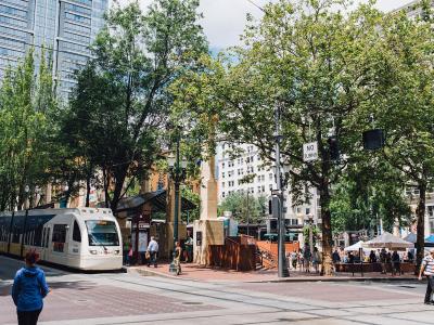 A street-level view of a city street corner showing a white MAX light rail train on the left, a transit shelter, trees, and pedestrians, with a "NO TURNS" traffic sign visible.