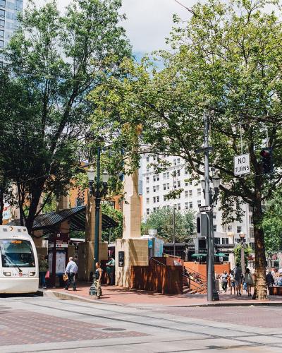 A street-level view of a city street corner showing a white MAX light rail train on the left, a transit shelter, trees, and pedestrians, with a "NO TURNS" traffic sign visible.