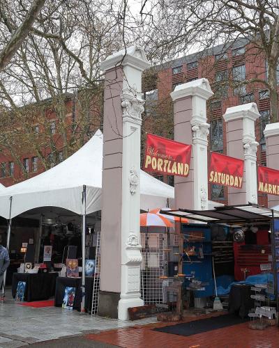 The image shows an outdoor market scene with white vendor tents, a large decorative archway structure, and people walking around; a banner on the archway reads "Welcome to Portland" and "Saturday Market".