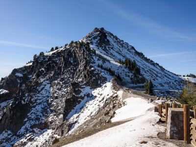 A view of a rugged, rocky mountain peak (Mount Scott) with snow patches, a winding paved road, and a wooden fence in the foreground, set against a blue sky.