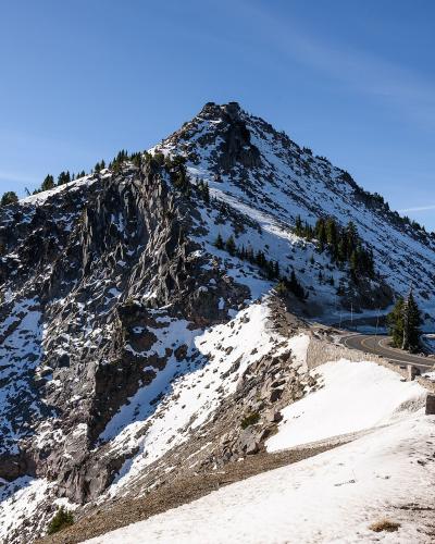 A view of a rugged, rocky mountain peak (Mount Scott) with snow patches, a winding paved road, and a wooden fence in the foreground, set against a blue sky.