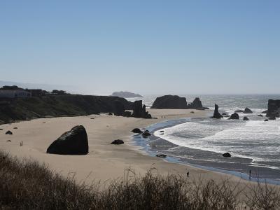 A coastal landscape showing a sandy beach with large sea stacks and rock formations, viewed from an elevated grassy bluff with a white building visible on the cliff to the left.
