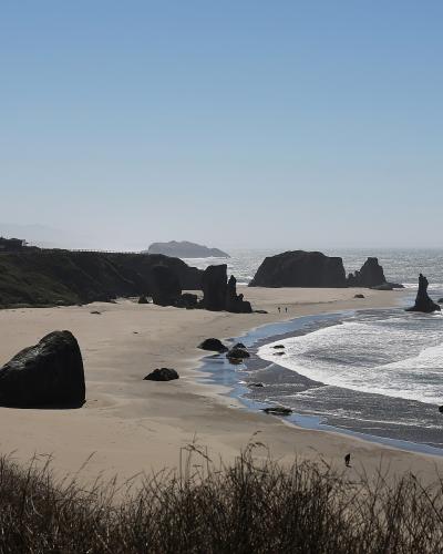 A coastal landscape showing a sandy beach with large sea stacks and rock formations, viewed from an elevated grassy bluff with a white building visible on the cliff to the left.