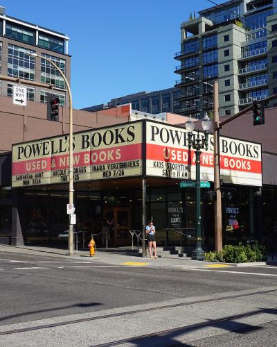 The image shows the exterior of a large bookstore with a prominent marquee sign reading "POWELL'S BOOKS" in large white letters, along with smaller text indicating "USED & NEW BOOKS" and event details; the building is situated on a city street corner