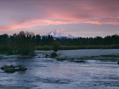 A landscape photograph showing a river in the foreground with a large, snow-capped stratovolcano (Mount Jefferson) in the background, framed by a treeline and a pink and purple sky.