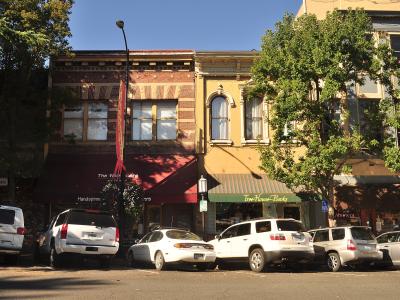A street-level view of a row of commercial buildings with storefronts, including one with a green awning labeled "Tree House Books" and another with a maroon awning, with several parked cars in the foreground.