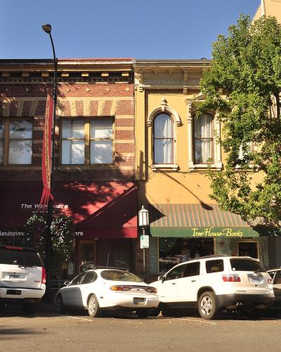 A street-level view of a row of commercial buildings with storefronts, including one with a green awning labeled "Tree House Books" and another with a maroon awning, with several parked cars in the foreground.