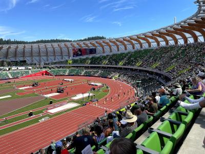 An outdoor athletics stadium featuring a distinctive curved wooden roof structure, green seating, and a red running track with athletes and spectators present.