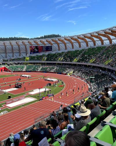 An outdoor athletics stadium featuring a distinctive curved wooden roof structure, green seating, and a red running track with athletes and spectators present.