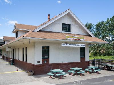 The image shows a white, single-story building with a brown shingled roof, identified by a sign as the "Mount Hood Railroad Depot," situated in a paved lot with green picnic tables.