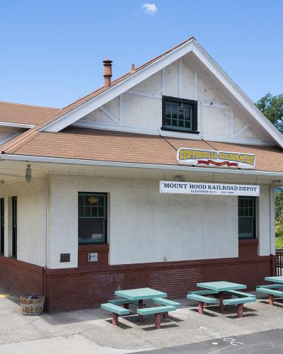 The image shows a white, single-story building with a brown shingled roof, identified by a sign as the "Mount Hood Railroad Depot," situated in a paved lot with green picnic tables.