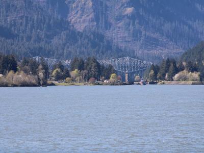 A long, white steel truss bridge spanning a wide river with forested mountains in the background and a small boat on the water.