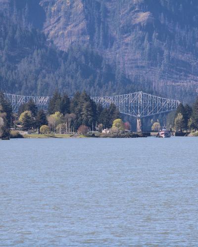 A long, white steel truss bridge spanning a wide river with forested mountains in the background and a small boat on the water.