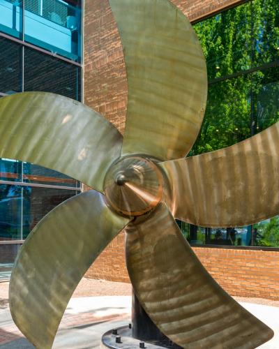 A large, five-bladed brass ship's propeller mounted on a pedestal outdoors in front of a modern building with glass walls and brick accents.