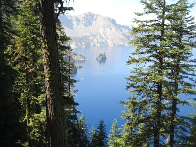 A scenic view of a deep blue crater lake surrounded by steep volcanic cliffs and evergreen trees, featuring a distinctive rocky island formation in the water.