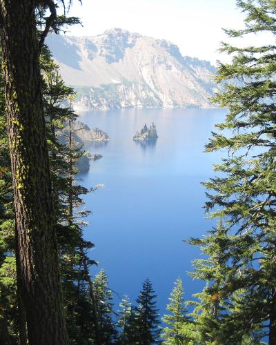 A scenic view of a deep blue crater lake surrounded by steep volcanic cliffs and evergreen trees, featuring a distinctive rocky island formation in the water.