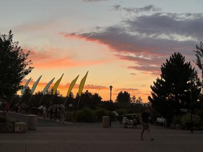 A paved plaza at dusk with people walking and sitting, featuring a row of tall flags (some yellow, some light blue/green) on the left and trees silhouetted against a sunset sky.
