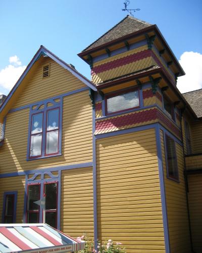 A yellow Victorian-style house with colorful trim, a tower, and a striped awning that reads "THE LATEST SCOOP FOR KIDS AND BABY".