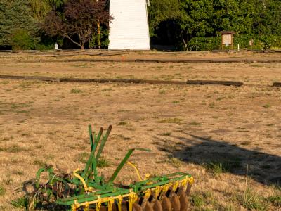 A rural landscape featuring a white wooden windmill tower and a vintage green disc harrow in a dry field.