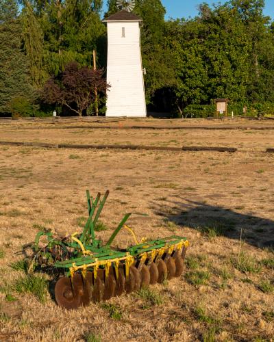 A rural landscape featuring a white wooden windmill tower and a vintage green disc harrow in a dry field.