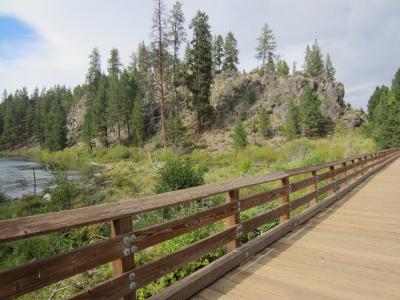 A wooden boardwalk with a brown railing running along the edge of a river, with a rocky, tree-covered hillside in the background.