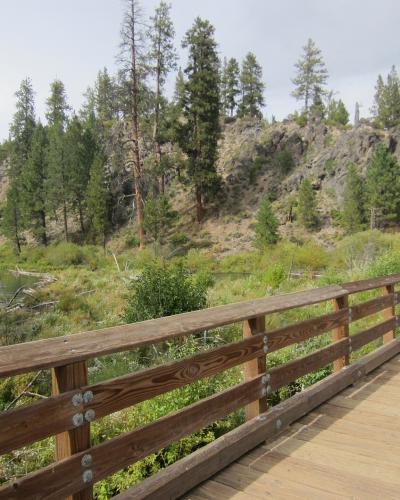 A wooden boardwalk with a brown railing running along the edge of a river, with a rocky, tree-covered hillside in the background.